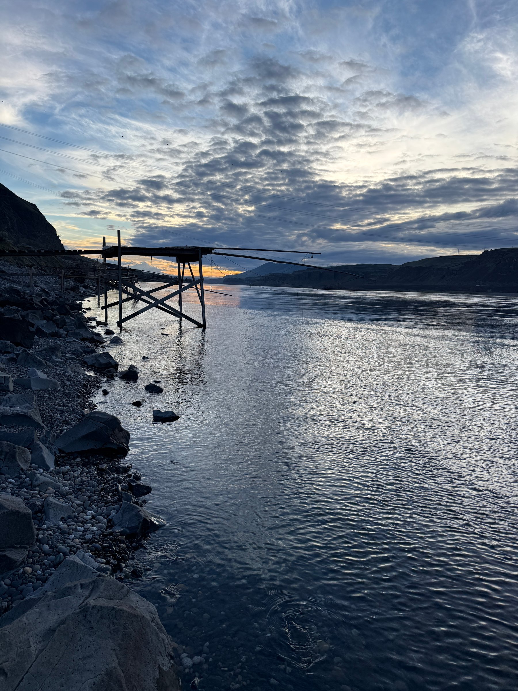 A wooden fishing platform with dip-net poles over a wide river at dusk, with basalt hills beyond.