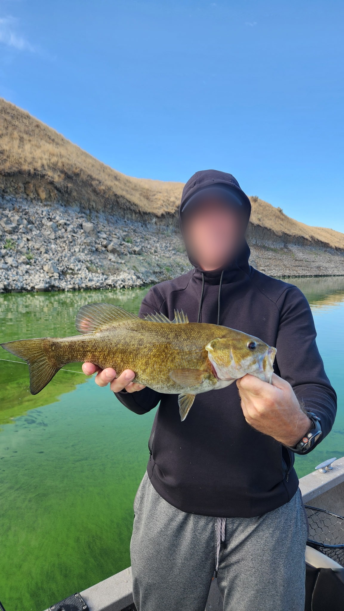 TA on the boat holding a smallmouth bass with both hands, face blurred, dry grass and a rocky bank behind