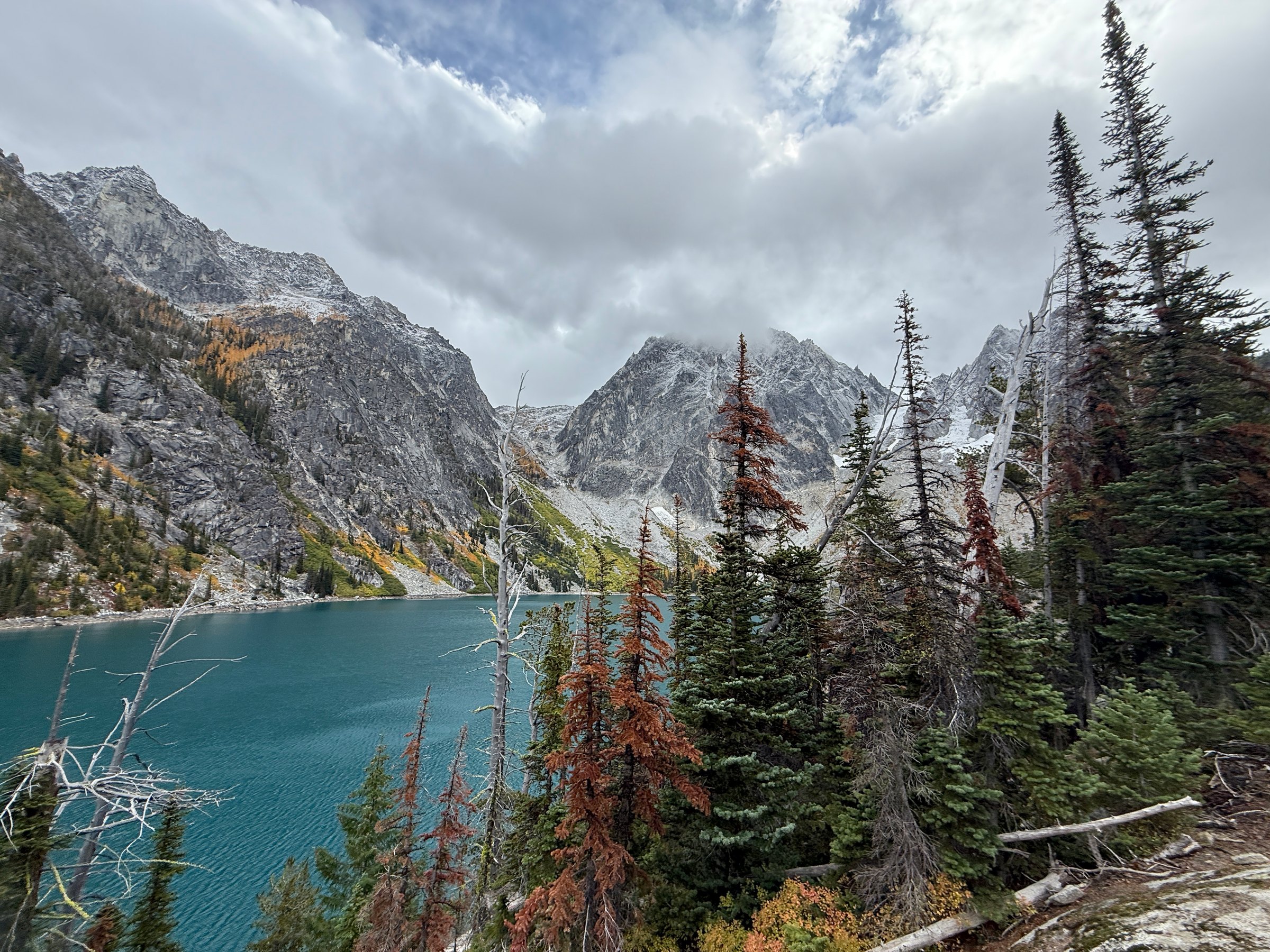 A turquoise alpine lake below snow-dusted peaks, with larches turning gold among the firs under a grey sky.