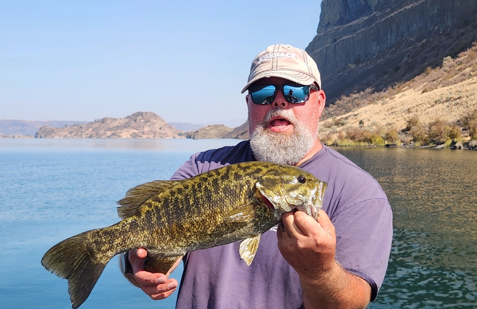 MD holding a smallmouth bass with both hands, basalt cliffs and dry slopes behind the water