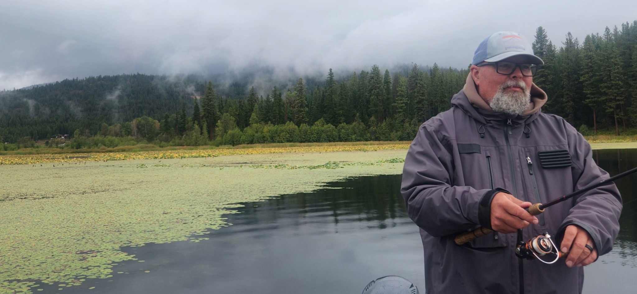 Mike holding a spinning rod on a boat, with lily pads, a misty forested hillside and low cloud behind.