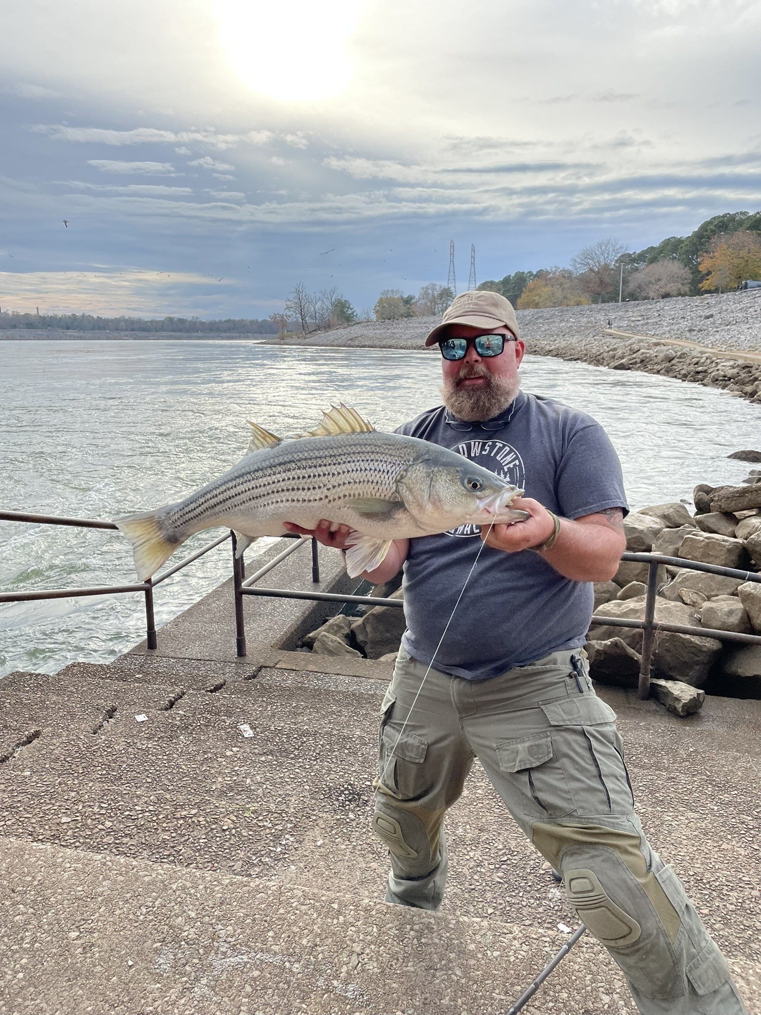 MD holding a striped bass on the concrete steps of a tailwater below a dam, the current behind him and riprap along the bank