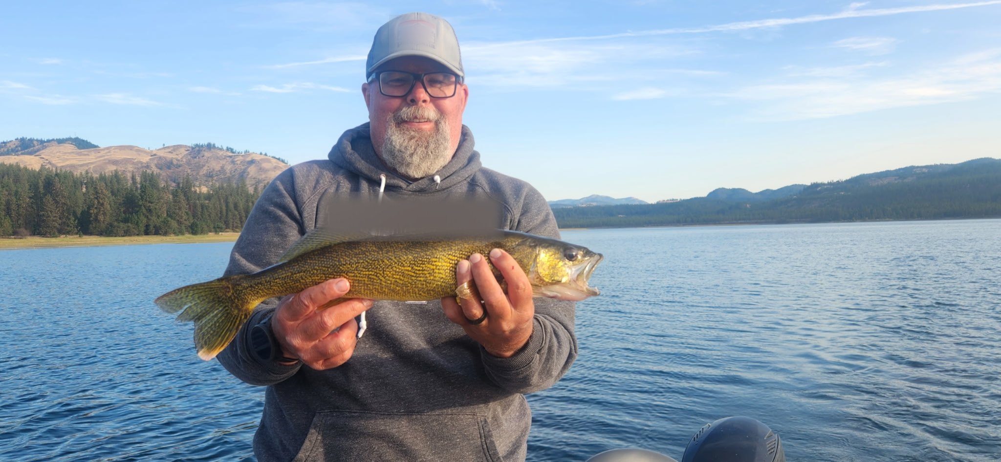 Mike holding a walleye on a boat, with dry hills and conifers behind in evening light.