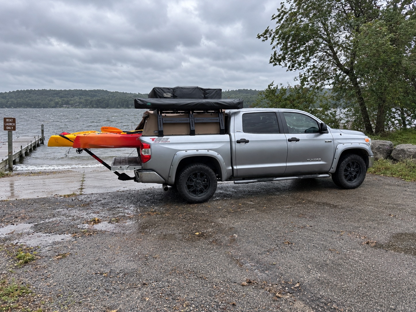 A silver pickup with two kayaks on a bed extender, parked at a boat launch under grey skies, with wind-chopped water behind it.