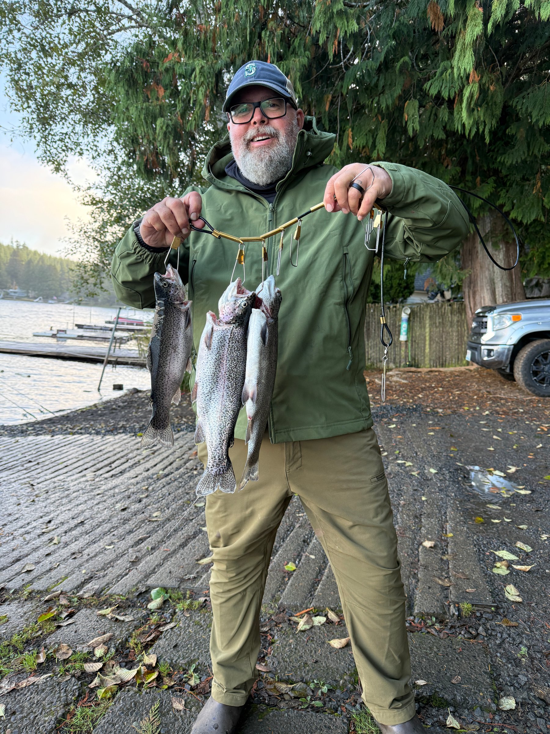 An angler in a green jacket and cap holds a stringer of three trout at the foot of a concrete boat ramp, with docks and the lake behind him.