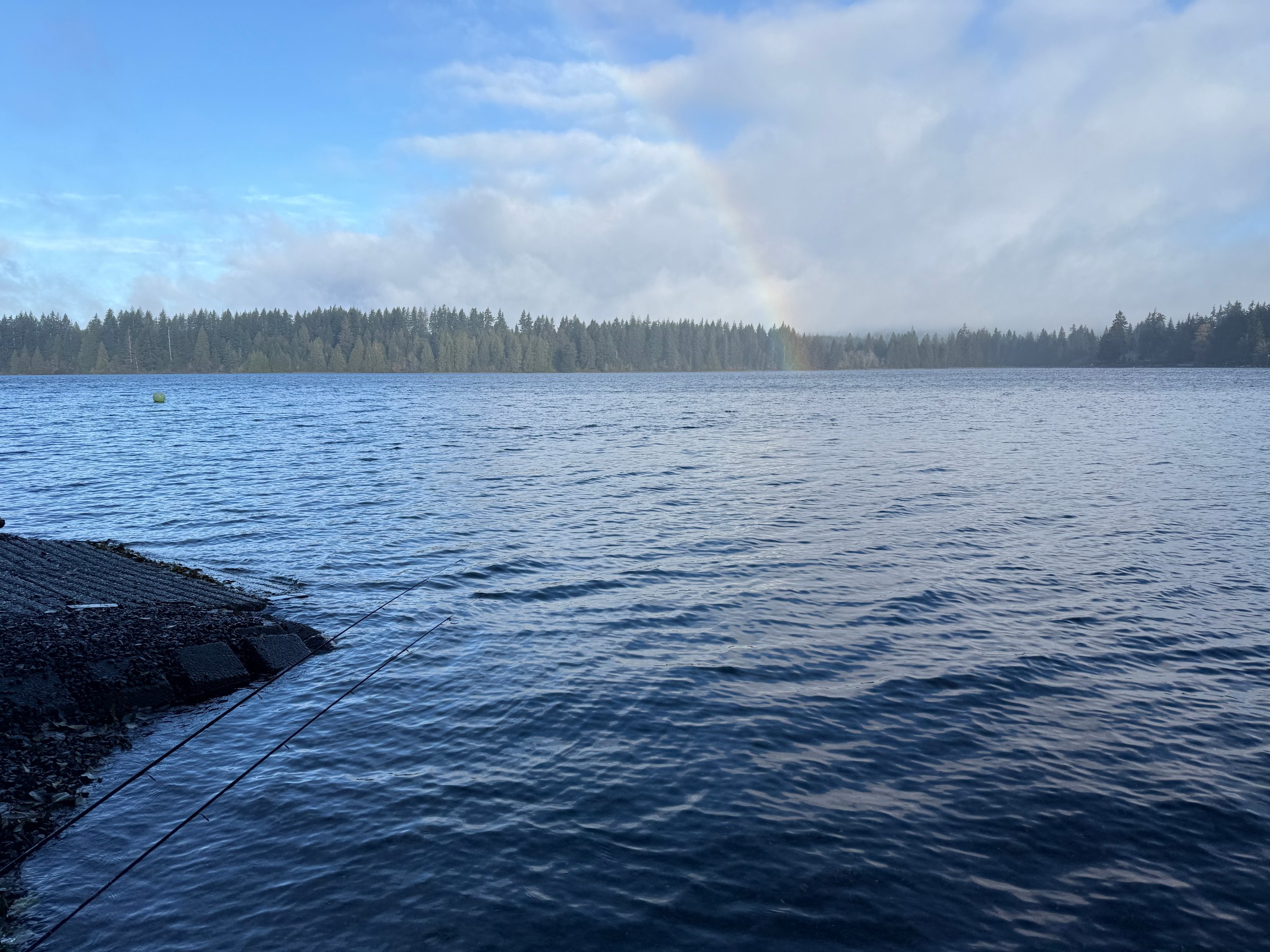 Two fishing rods angled over rippled blue water beside a concrete boat ramp, with a rainbow dropping into a tree-lined far shore under breaking cloud.